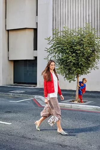 Fotoproduktion Superwoman Eine Brünette Frau mit roter Jacke, High Heels und Handtasche geht auf der Straße entlang, und wird von einem Kind in Superman-Kostüm beobachtet