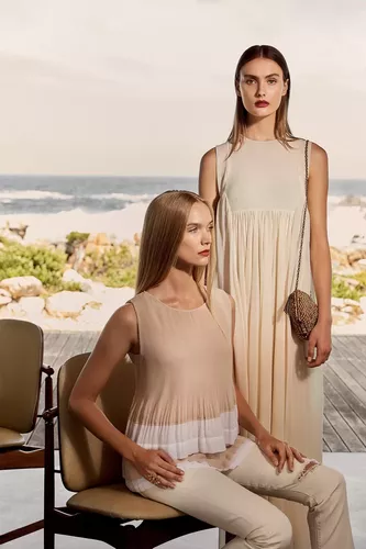 Fotoproduktion Beige Beach Ein Modell sitzt auf einem Stuhl, das andere Modell steht daneben und guckt in die Kamera im Hintergrund Strand